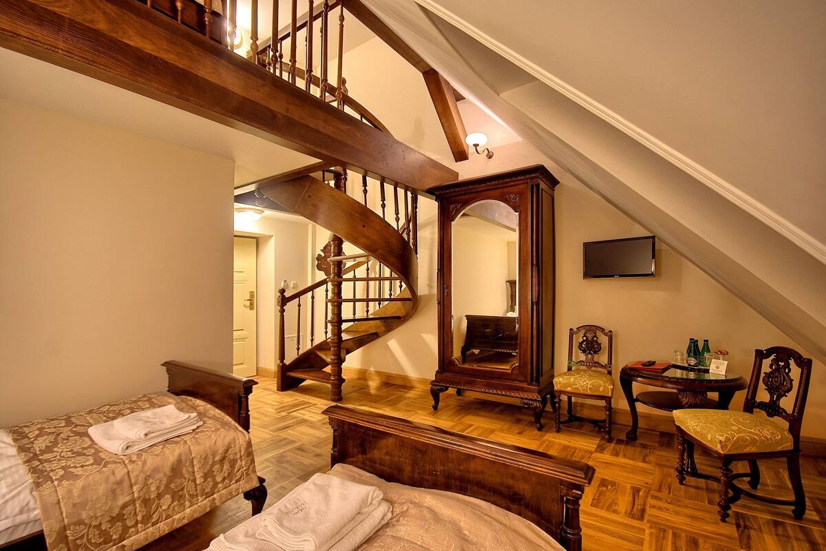 Um quarto com escada de madeira, uma cama com colcha florida, uma cadeira e uma pequena mesa com garrafas.
