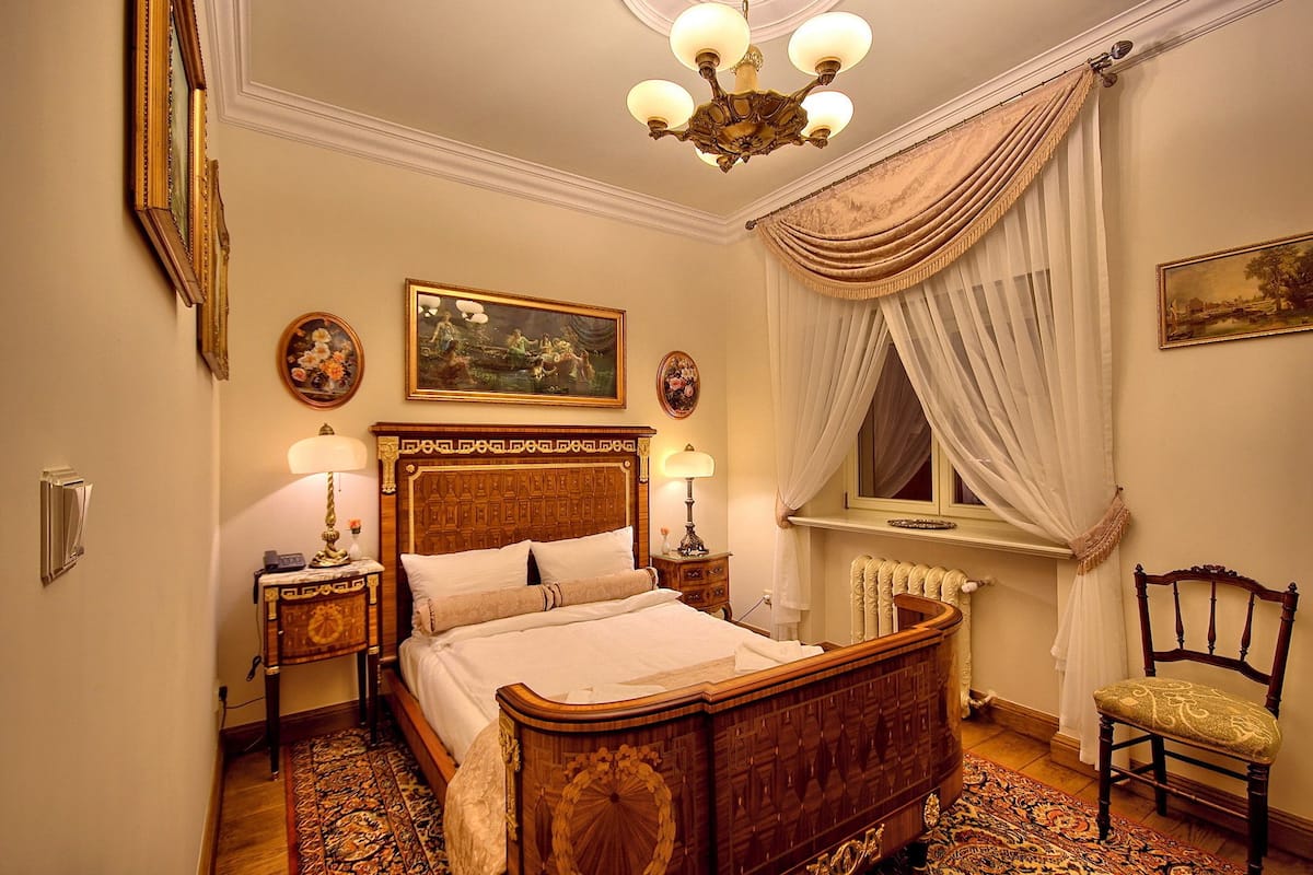 Um quarto tradicional com uma cama de madeira, duas mesas de cabeceira, um candelabro e uma janela com cortinas.