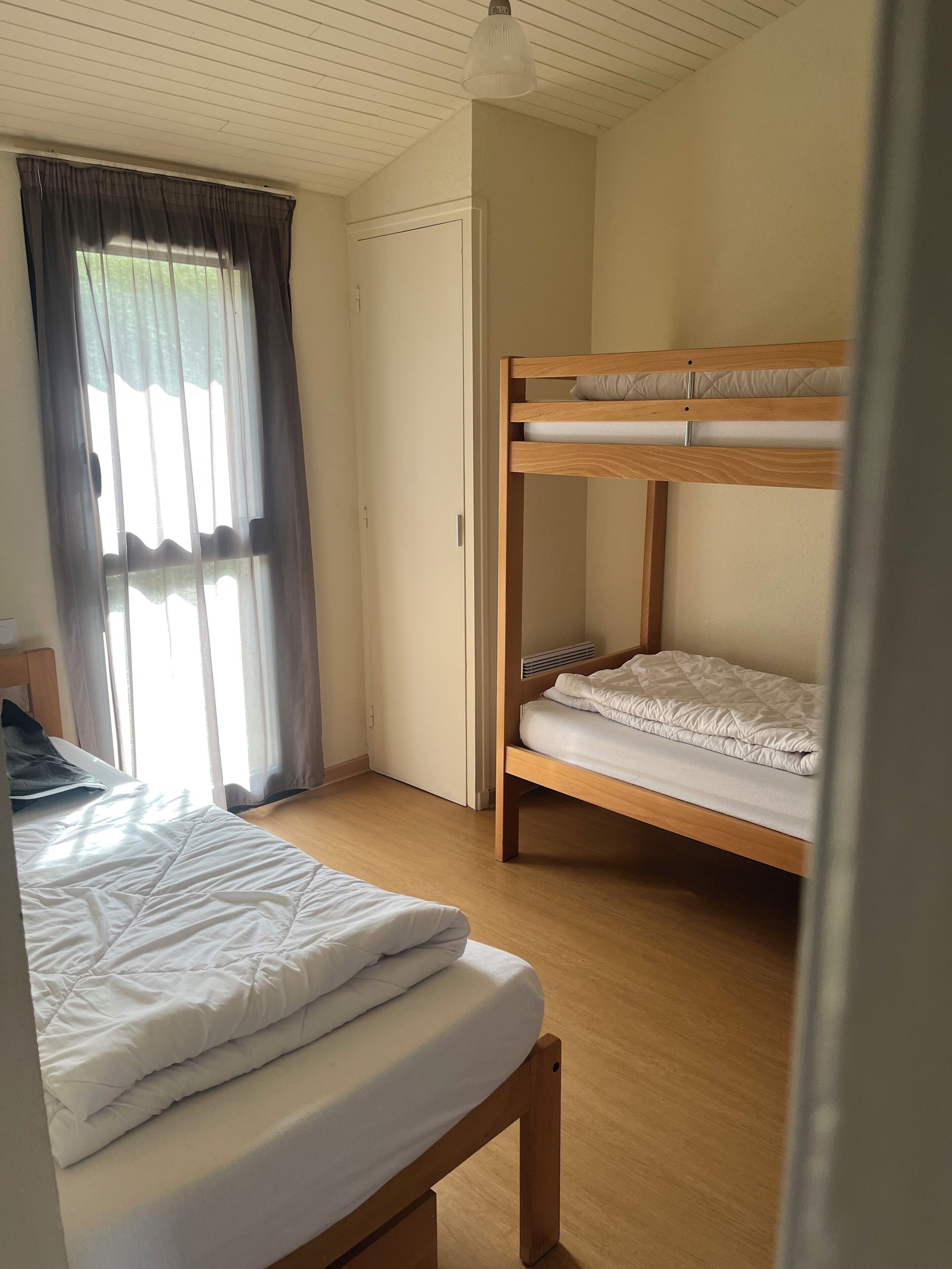 A room with a bunk bed, a single bed, a window with curtains, and a door.