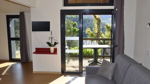 A modern living room with a grey sofa, a red shelf with a potted plant, and a sliding glass door leading to an outdoor patio with a table and chairs. - Domaine Aigoual Cévennes (Meyrueis)