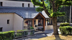 A building with a sloped roof, a wooden porch, and a small garden area with a bench. - Domaine Aigoual Cévennes (Meyrueis)