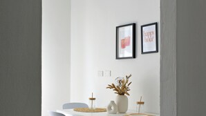 Un coin repas avec une table et des chaises blanches, un vase contenant des plantes séchées et deux tableaux encadrés accrochés au mur.