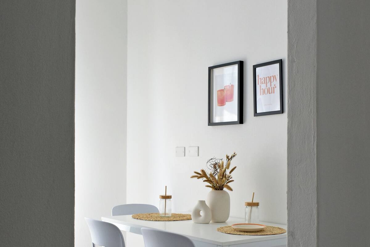 Ein Essbereich mit einem weißen Tisch und Stühlen, einer Vase mit Trockenpflanzen und zwei gerahmten Bildern an der Wand.