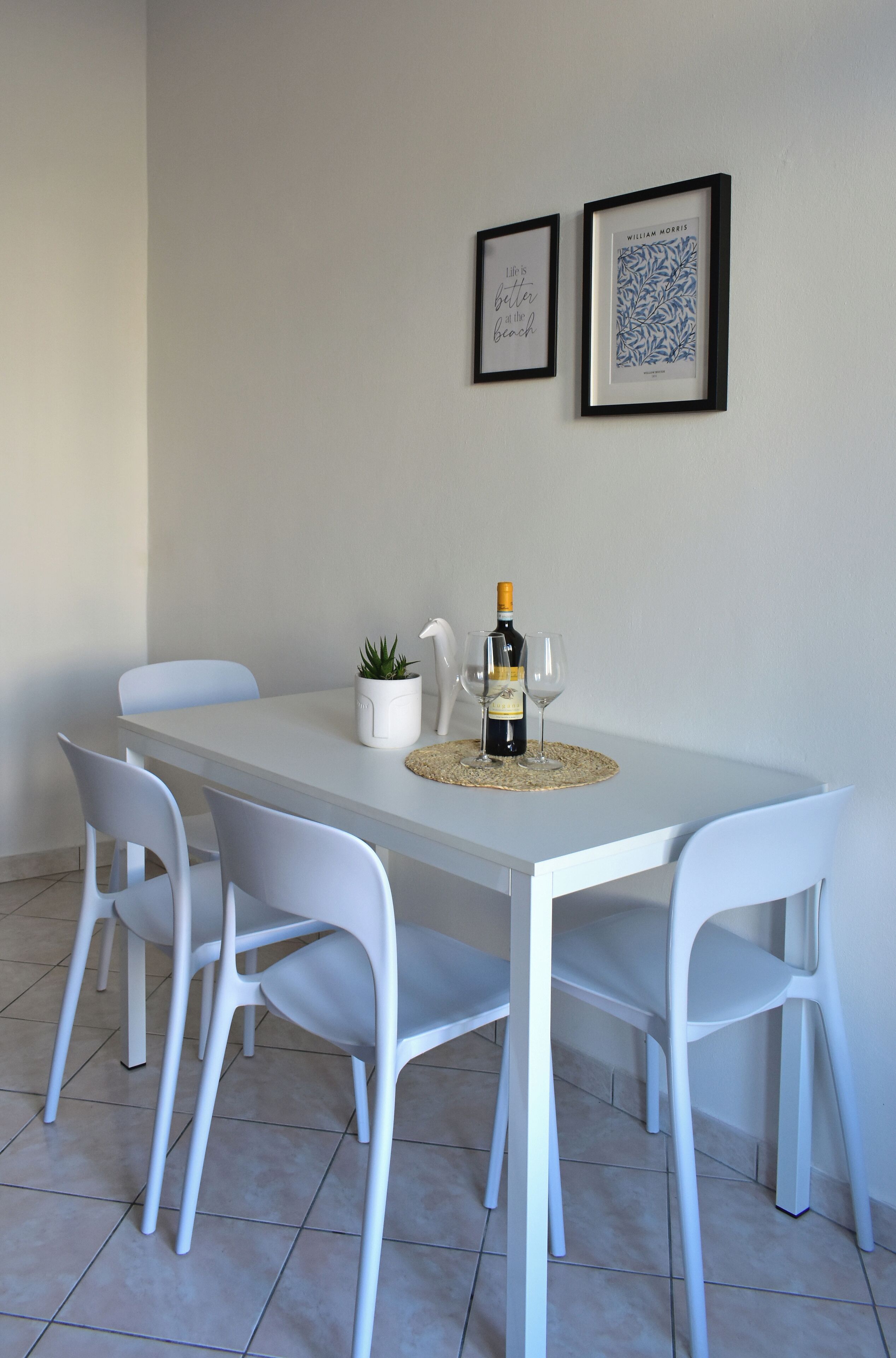 Ein Esstisch mit vier weißen Stühlen, einer Weinflasche, zwei Weingläsern und einem kleinen Topfpflanzchen. Über dem Tisch hängen zwei gerahmte Bilder an der Wand.