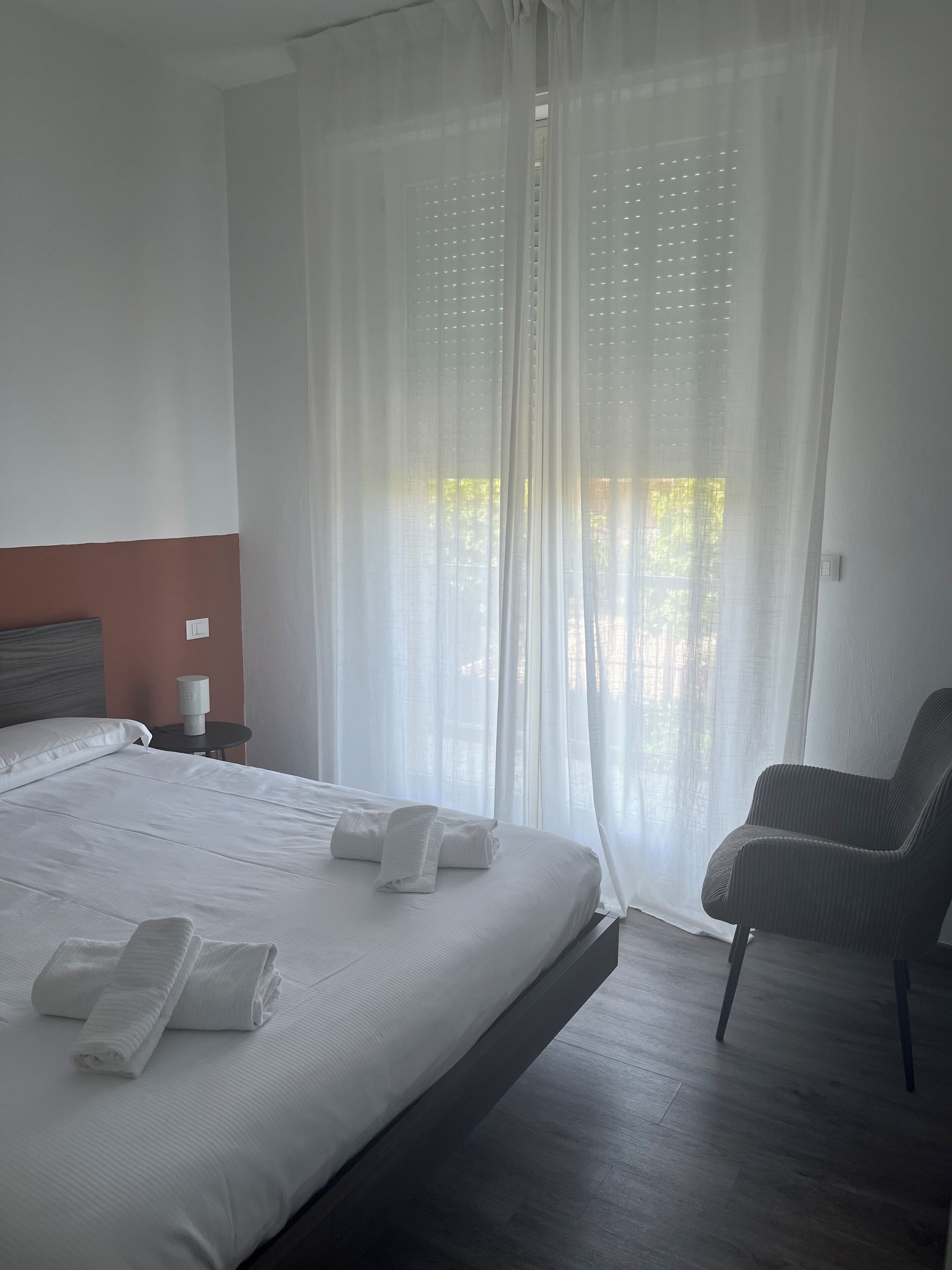 Una habitación de hotel con una cama grande, una silla, una mesita de noche con lámpara y cortinas translúcidas.
