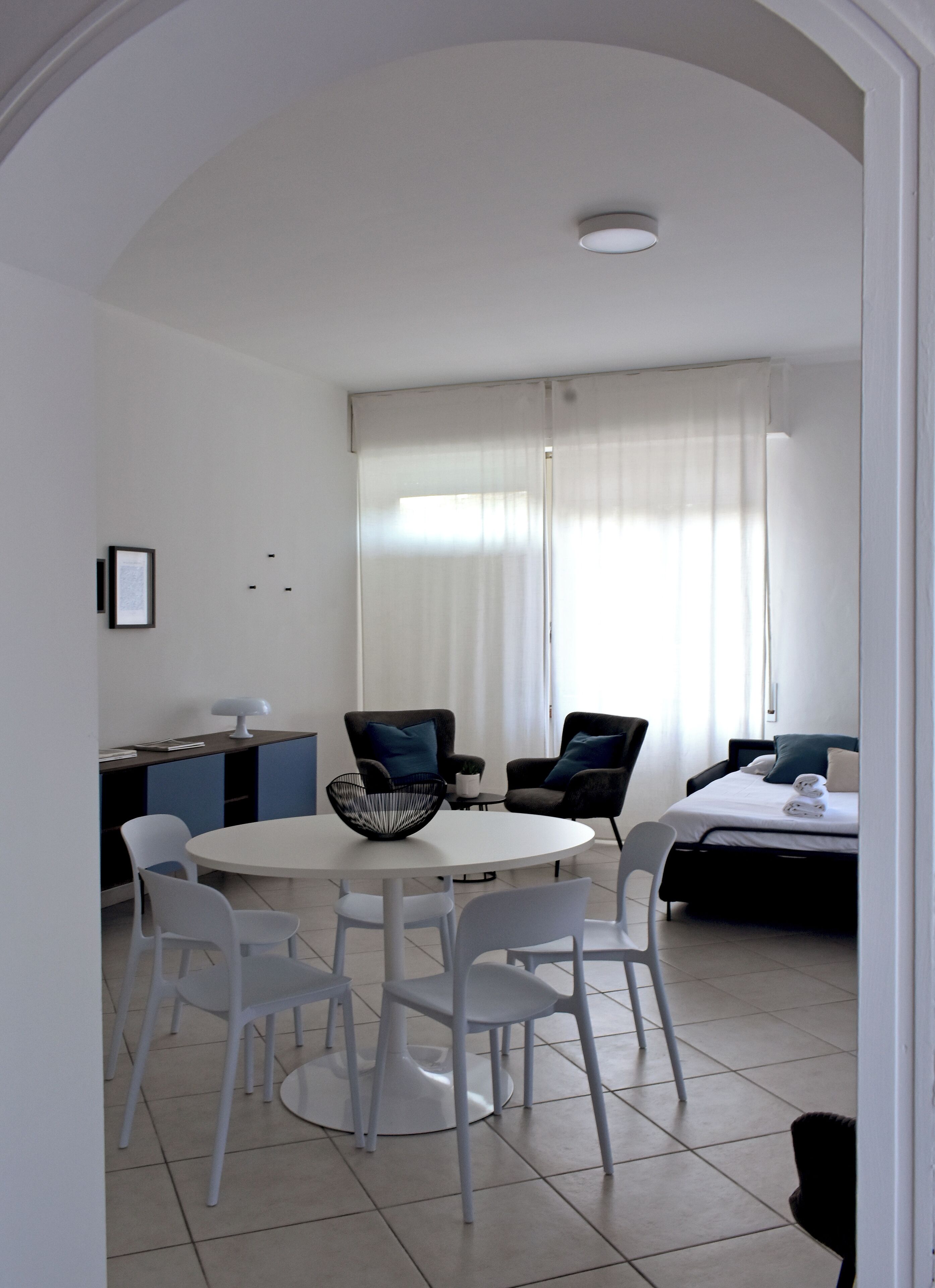 A modern, well-lit room with a round dining table surrounded by white chairs, a small side table with a bowl, and a bed with a dark headboard and pillows.