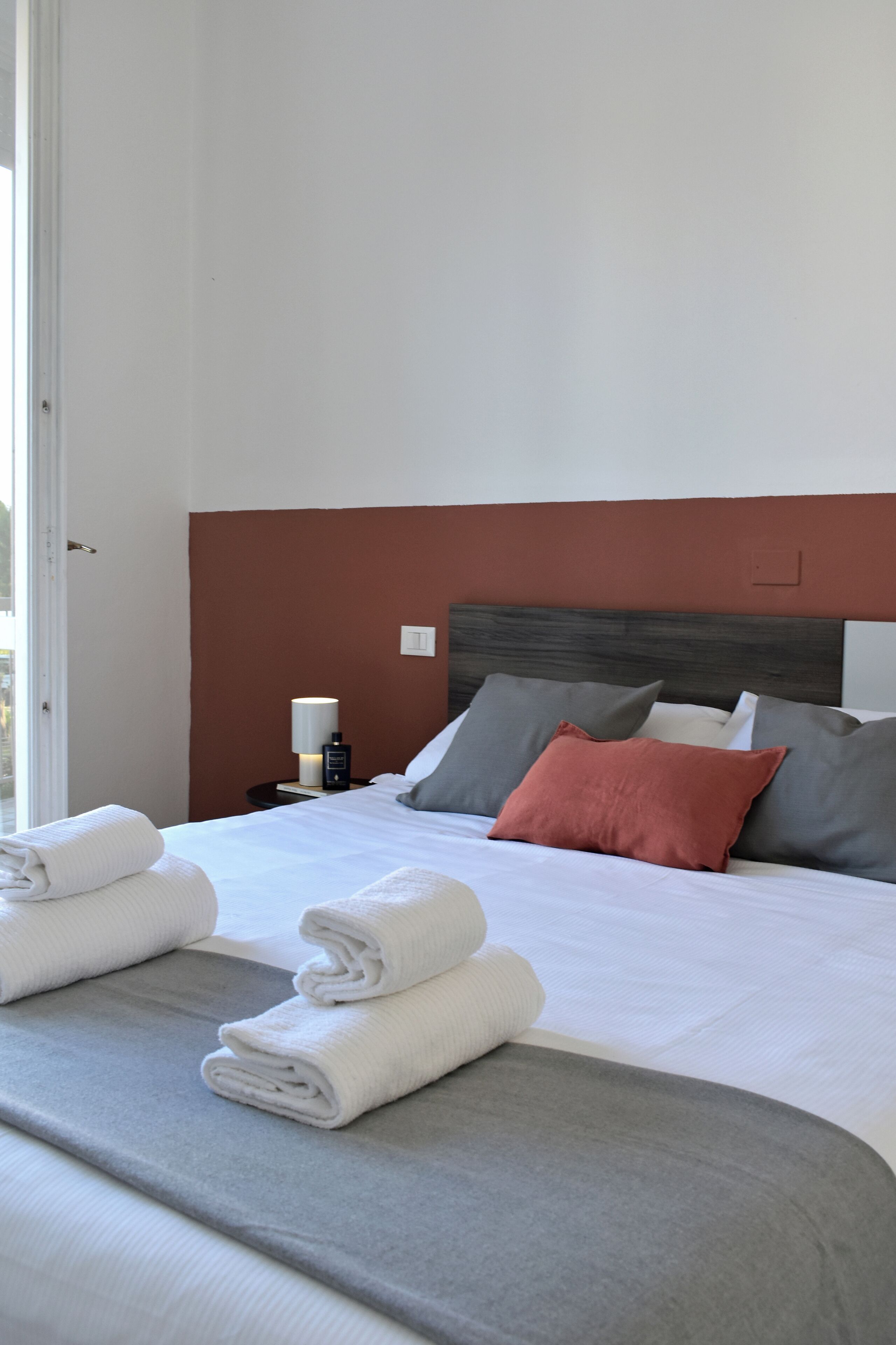 A neatly made bed with white linens and rolled towels, a bedside table with a lamp, and a brown headboard.