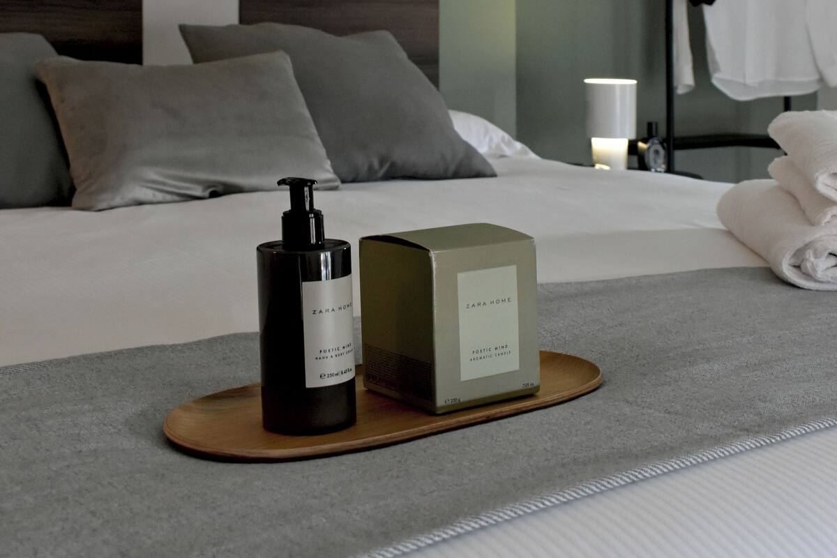 Ein ordentlich bezogenes Bett mit Kissen, eine Parfumflasche und eine Kiste von Zara Home auf einem Tablett.