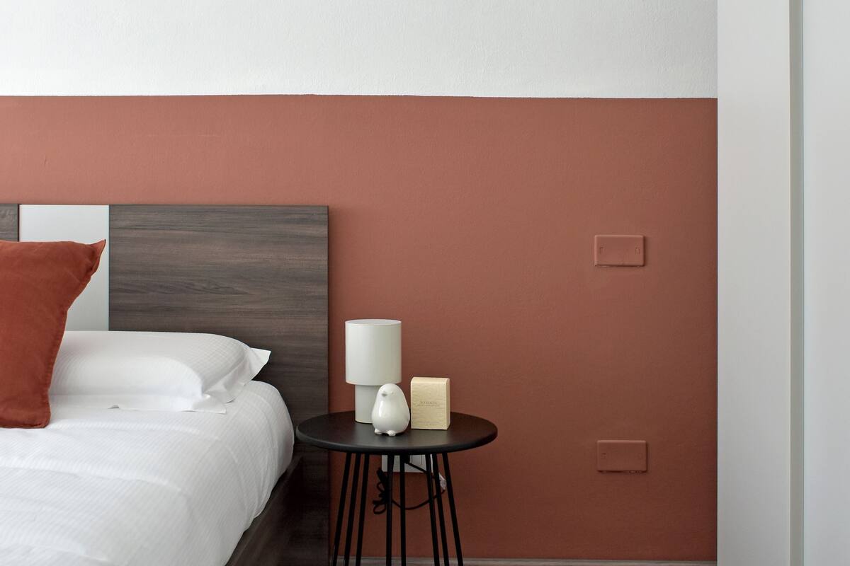 Ein Hotelzimmer mit einem Bett, einem kleinen runden Tisch mit Lampe und einer Schachtel, sowie einer Wand mit einem Lichtschalter.