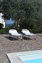 Two sun loungers with white covers, a stone-paved area, and a pool edge.