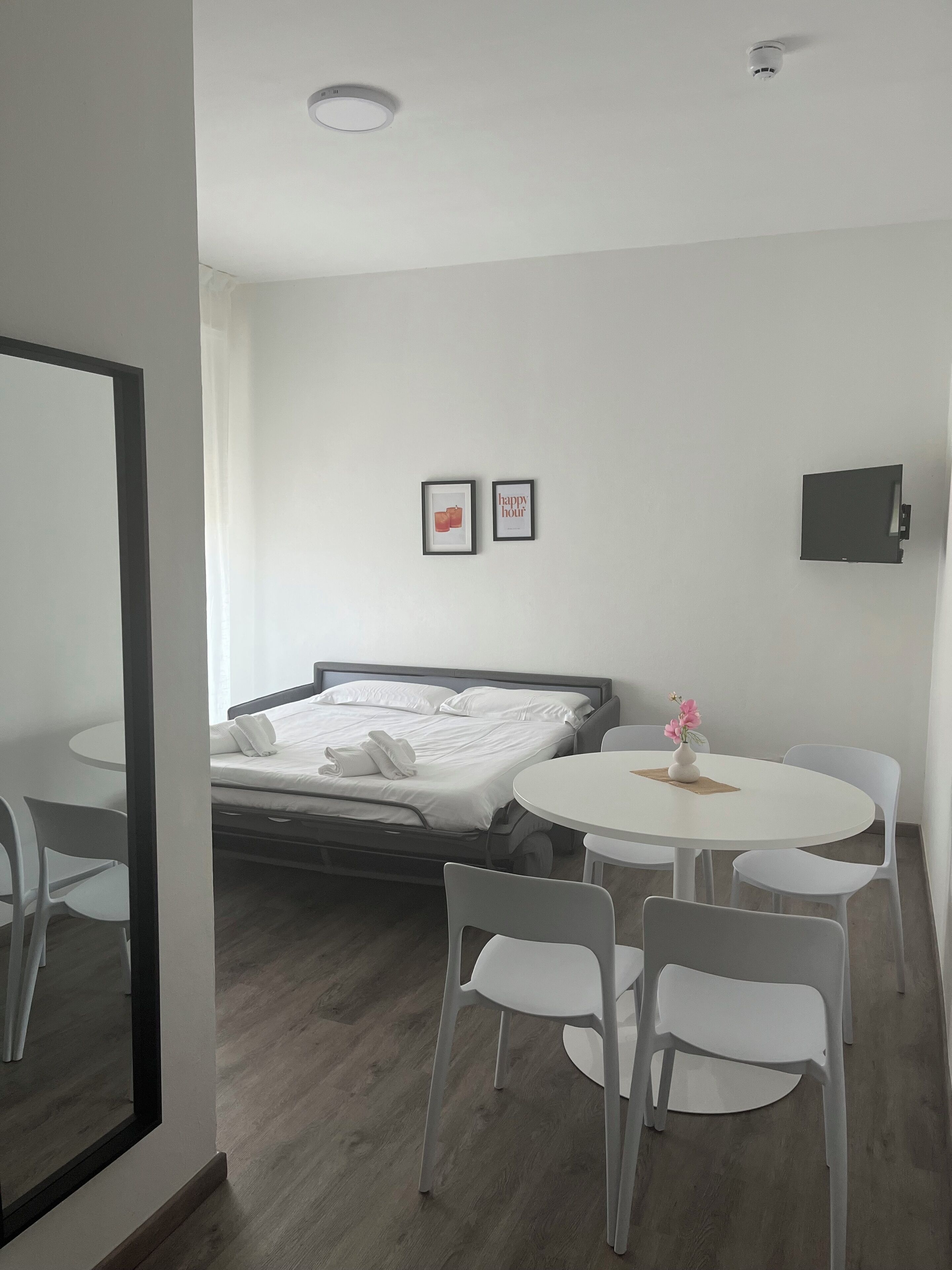 A modern hotel room with a bed, a round dining table, white chairs, a small table with a vase, and two framed pictures on the wall.