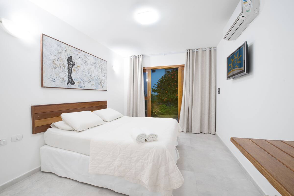 Une chambre à coucher avec un lit, une tête de lit en bois, un grand tableau, un climatiseur et une vue sur l’extérieur.