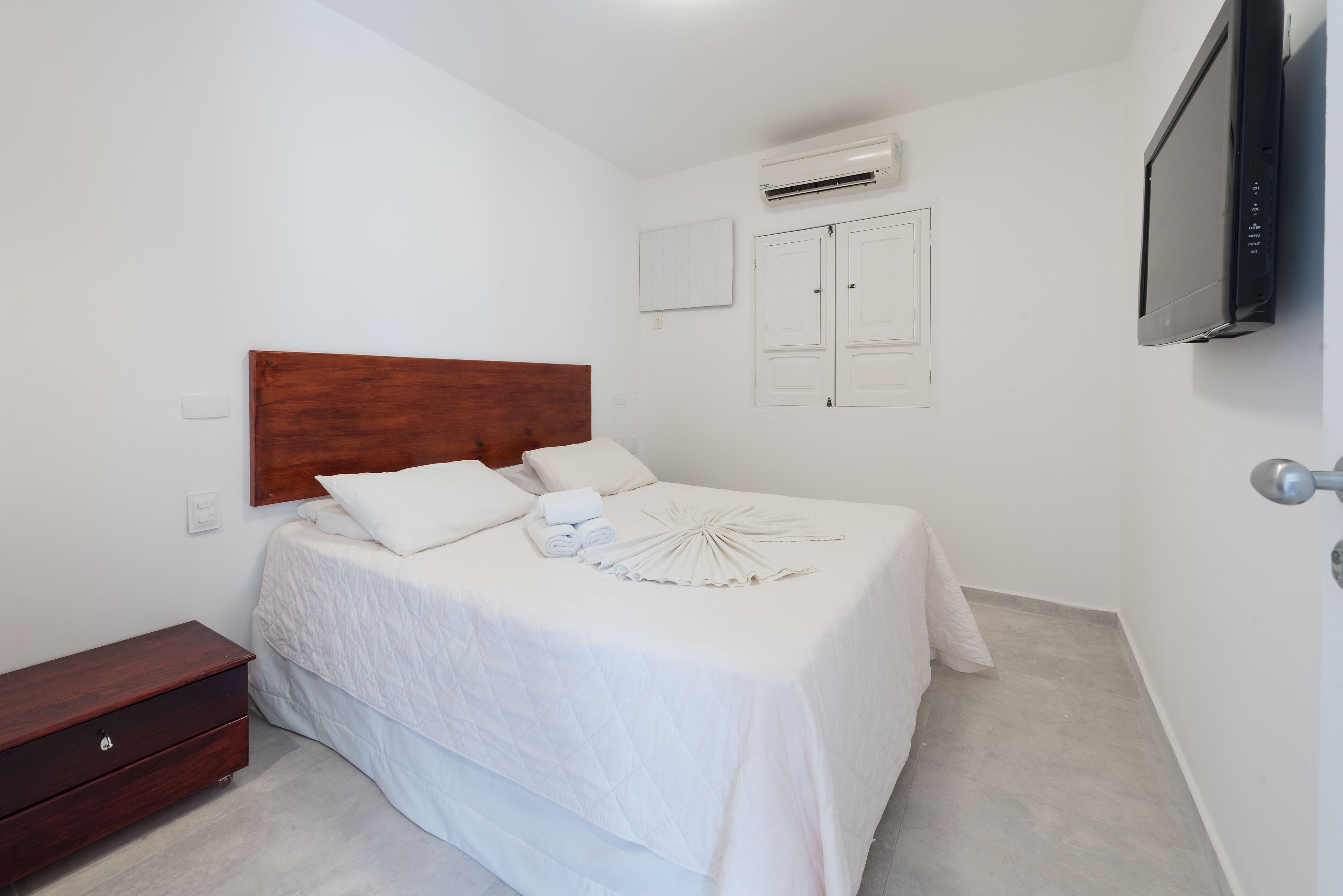 A bedroom with a wooden headboard, a bed with white bedding, a bedside table, a wardrobe, an air conditioning unit, and a television mounted on the wall.