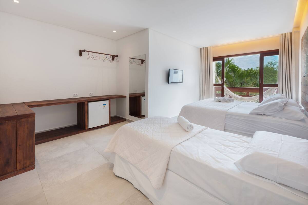 Une chambre d’hôtel avec deux lits, un bureau, un miroir, une télévision et une grande fenêtre donnant sur des palmiers.