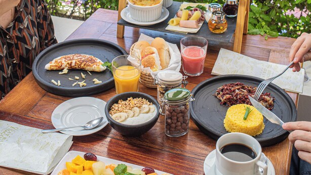 A dining table with breakfast items, a pool, and greenery in the background. - Pousada Quatro Estações (Ipojuca)