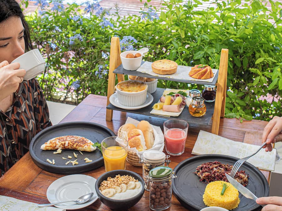 Una mesa de comedor con alimentos para el desayuno, una piscina y follaje al fondo.