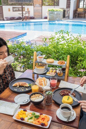 A dining table with breakfast items, a pool in the background, and a person drinking from a cup. - Pousada Quatro Estações (Ipojuca)
