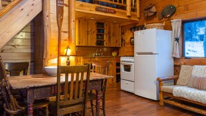 A wooden cabin interior with a dining table, chairs, a kitchen with a stove and refrigerator, a wooden staircase, and a window with a view of the outside. - Pioneer Guest Cabins (Crested Butte)