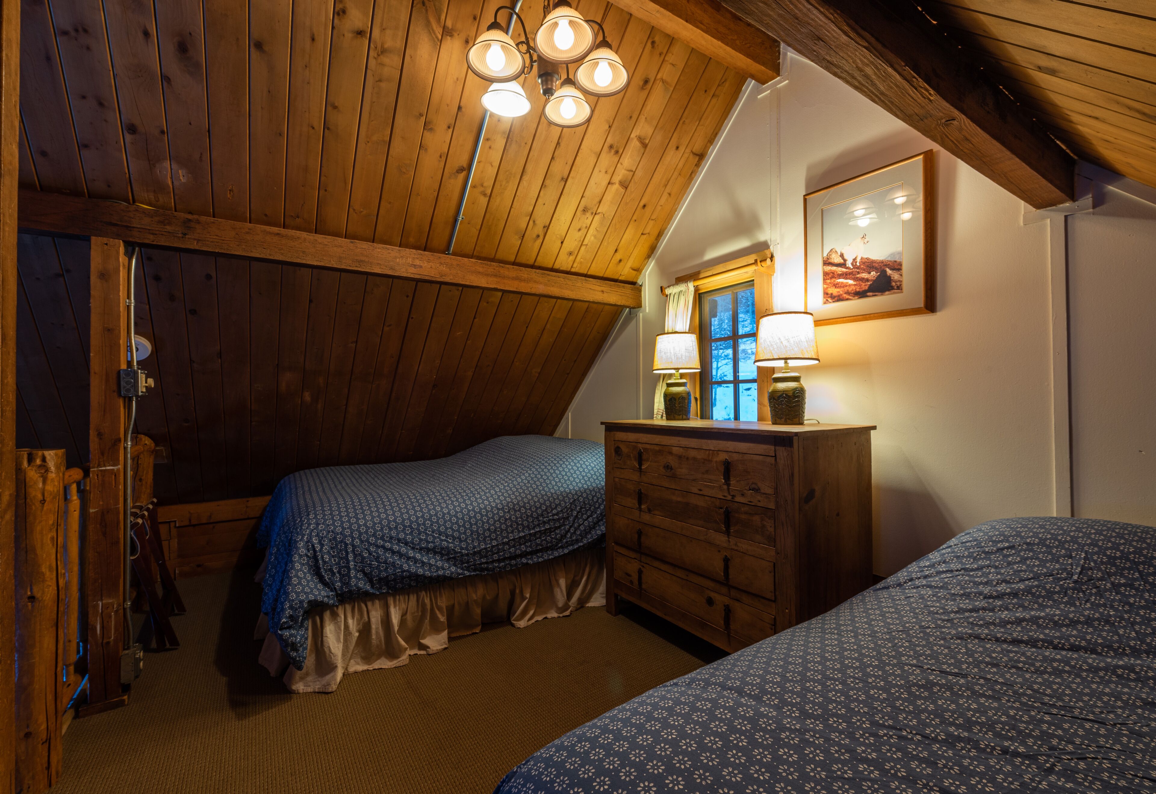A bedroom with two beds, wooden ceiling beams, a dresser, and a window with curtains.