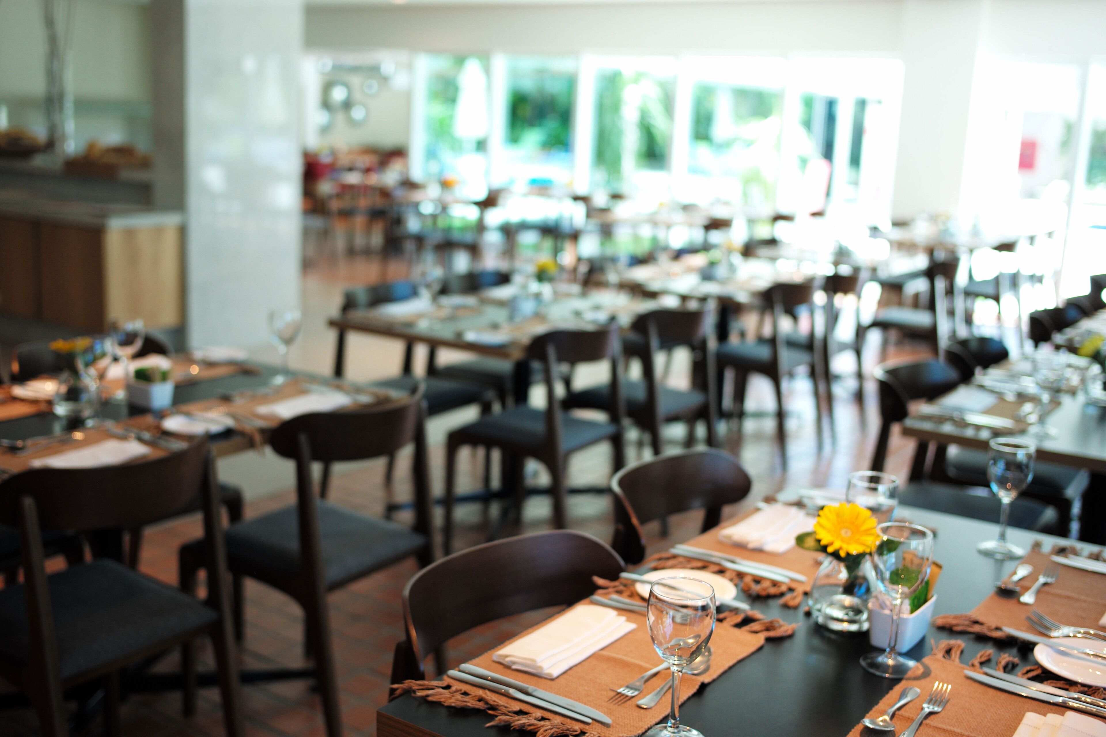Un restaurante con varias mesas dispuestas para comer, sillas y un jarrón decorativo central con una flor amarilla.