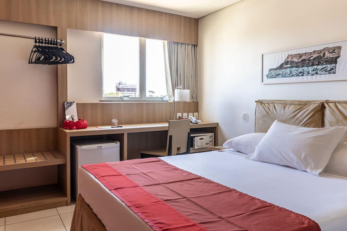 Habitación de hotel con cama, escritorio, silla, kitchenette pequeño y ventana con cortinas.
