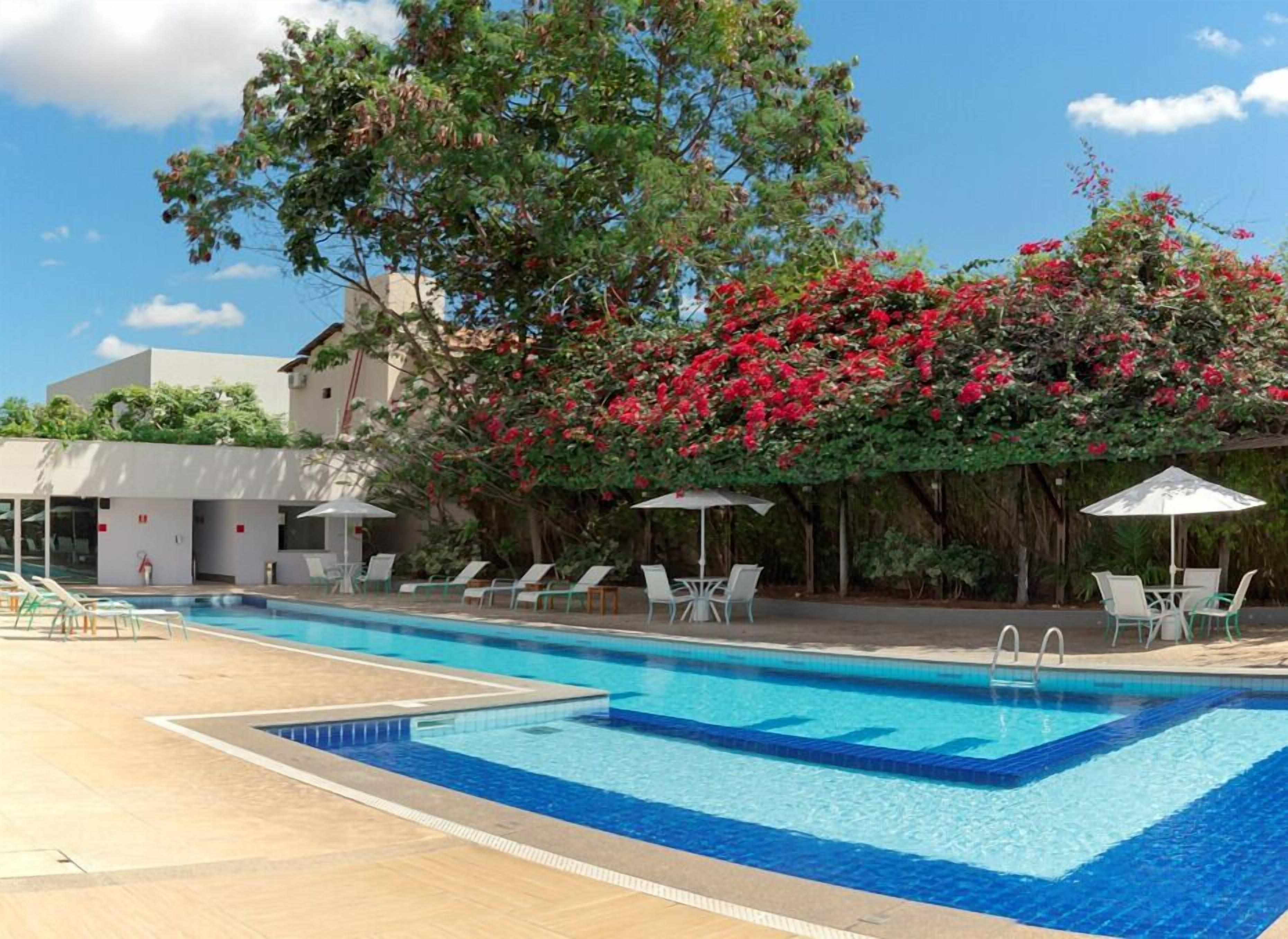 Piscina con sillas de descanso y sombrillas, rodeada de exuberante vegetación y un edificio.