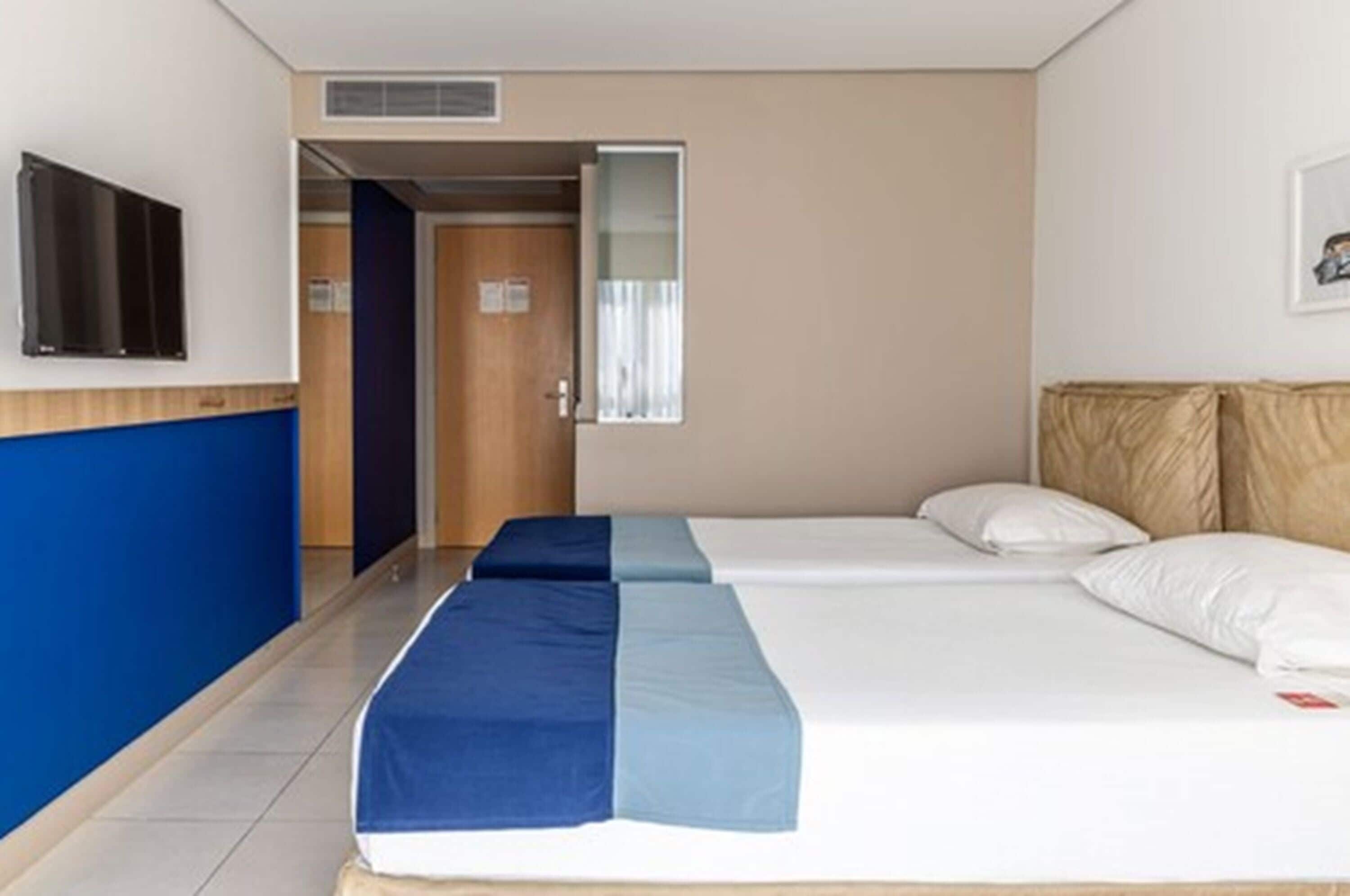 Een hotelkamer met twee bedden, een aan de muur bevestigde televisie en een deur naar een andere kamer.