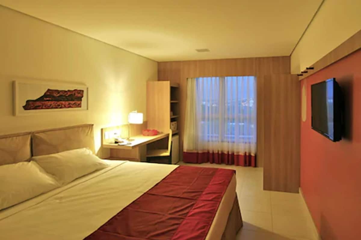 Una habitación de hotel con cama, escritorio, televisión y ventana con cortinas.