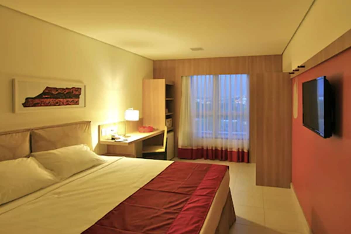 Una habitación de hotel con una cama, un escritorio con lámpara, televisión y ventana con cortinas.