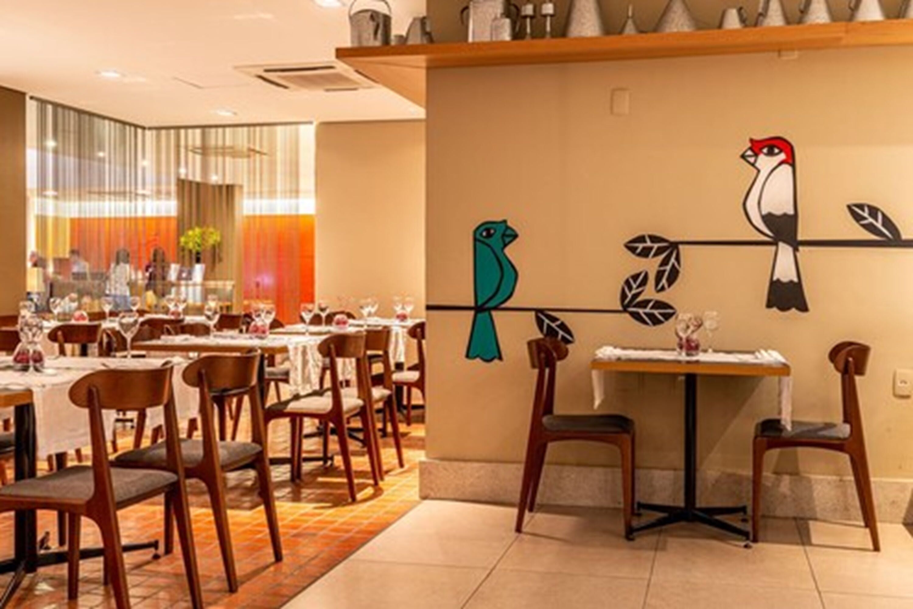 Interior de restaurante con sillas y mesas de madera, un mural de aves en la pared y una paleta de colores cálidos.