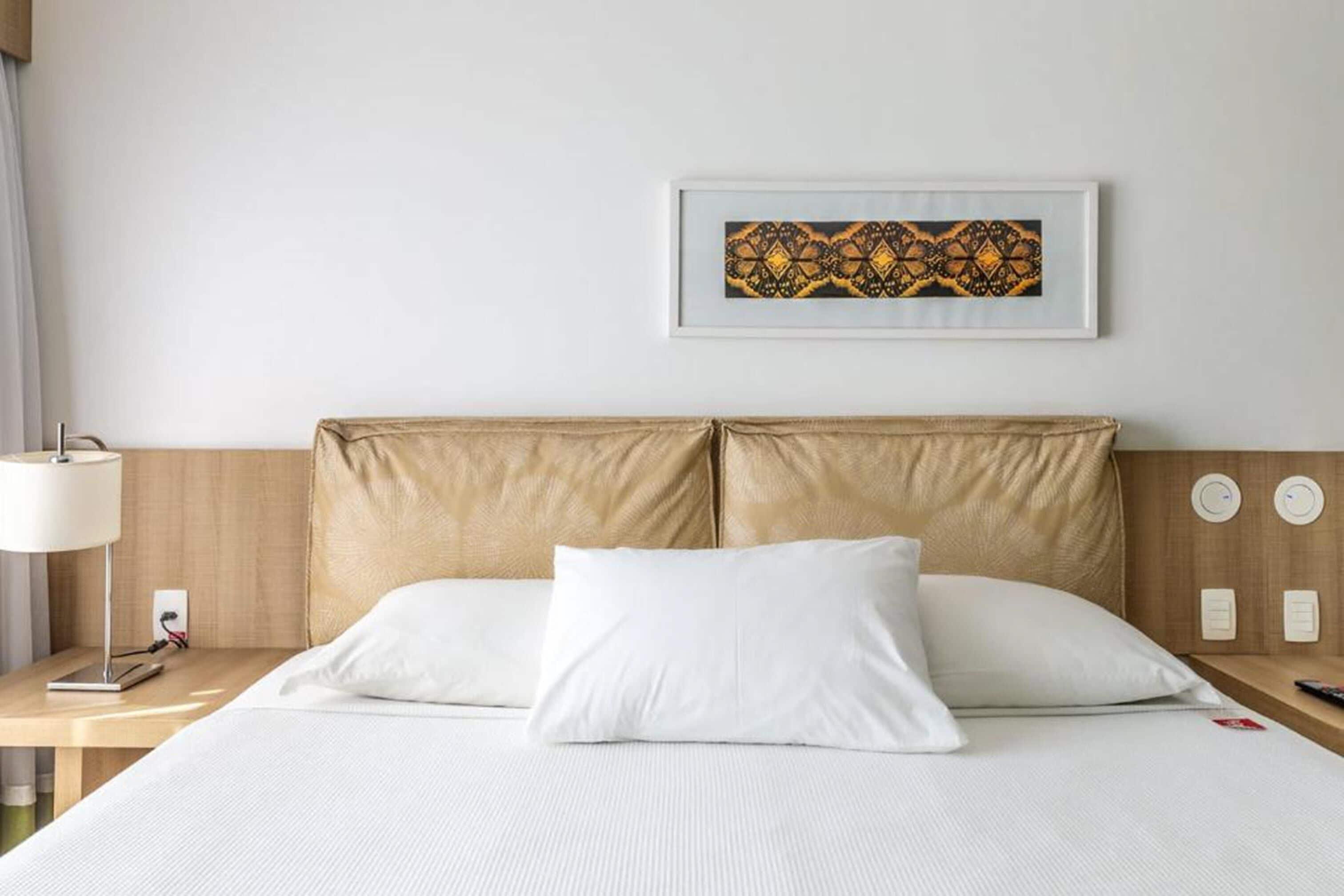Ein Hotelzimmer mit Bett, weißen Kissen, einem Nachttisch mit Lampe, einem gerahmten Kunstwerk an der Wand und einem hölzernen Kopfteil.
