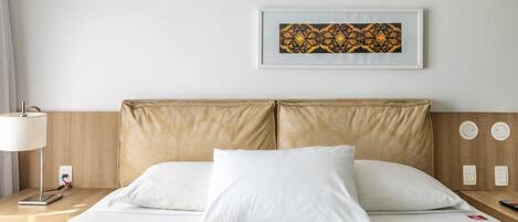 Ein Hotelzimmer mit Bett, weißen Kissen, einem Nachttisch mit Lampe, einem gerahmten Kunstwerk an der Wand und einem hölzernen Kopfteil.