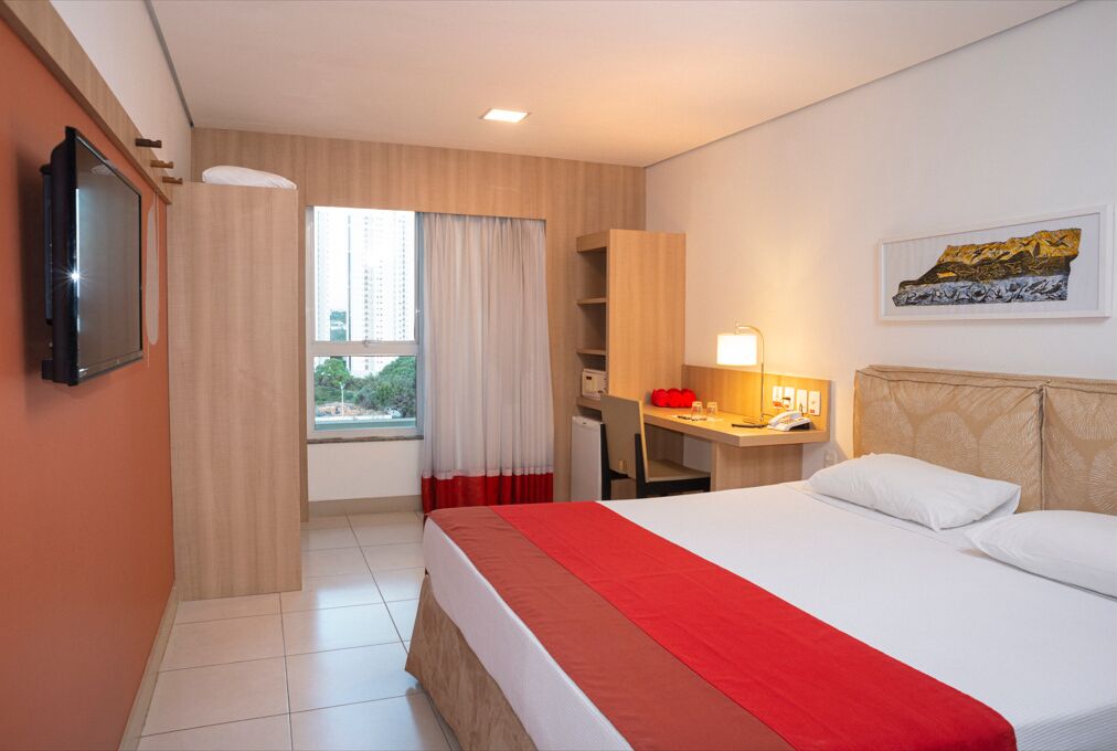 Een hotelkamer met een bed, een bureau, een televisie, een raam met gordijnen en een schilderij aan de muur.