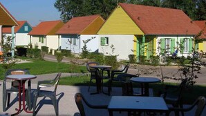 A row of colorful cottages with red roofs and green shutters, outdoor seating with plastic chairs and tables, and a grassy area with trees in the background. - Résidence Marina Holyder (Giffaumont-Champaubert)