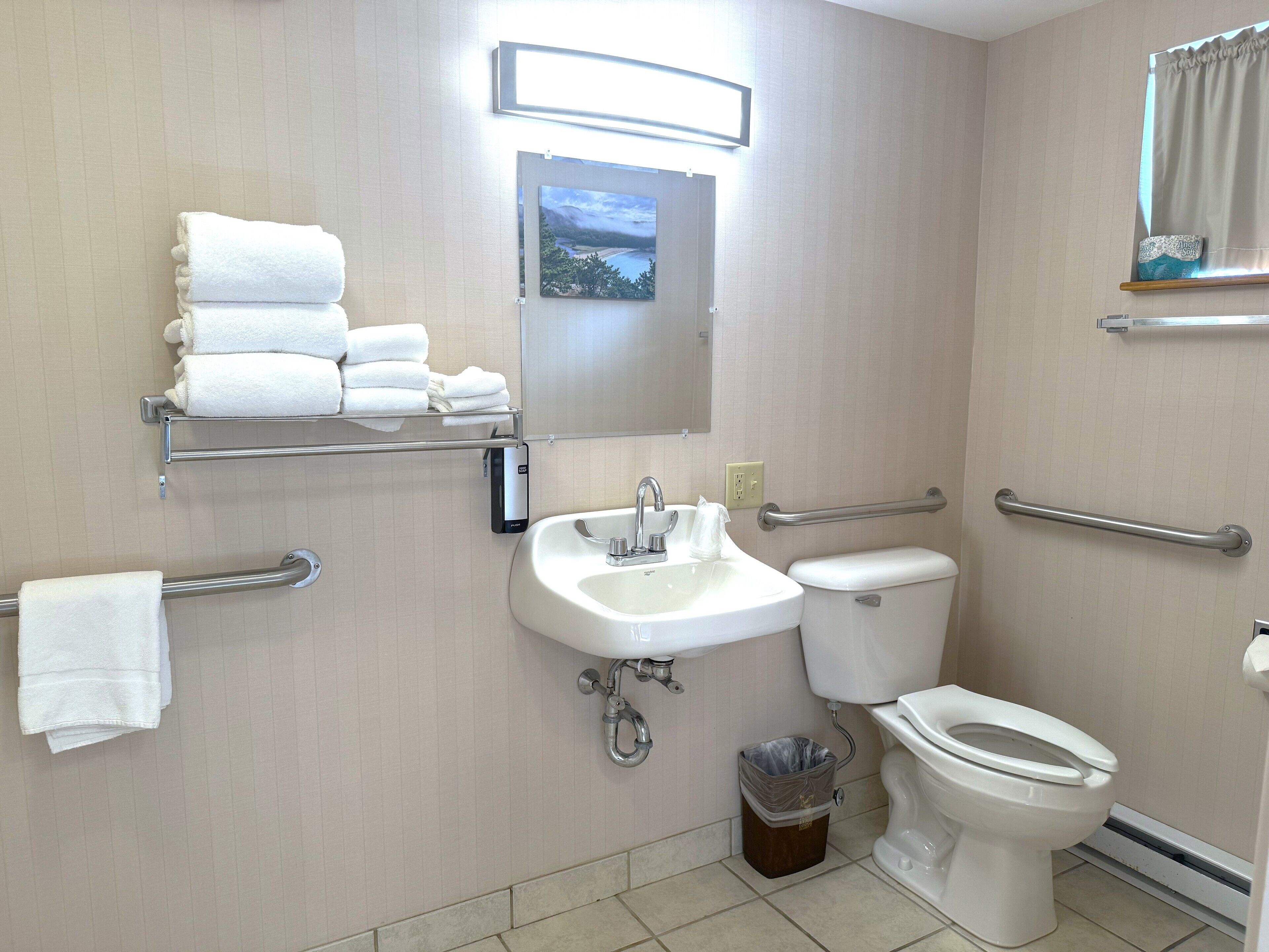 A bathroom with a sink, toilet, and handrails. There are towels stacked on a shelf and a trash can.