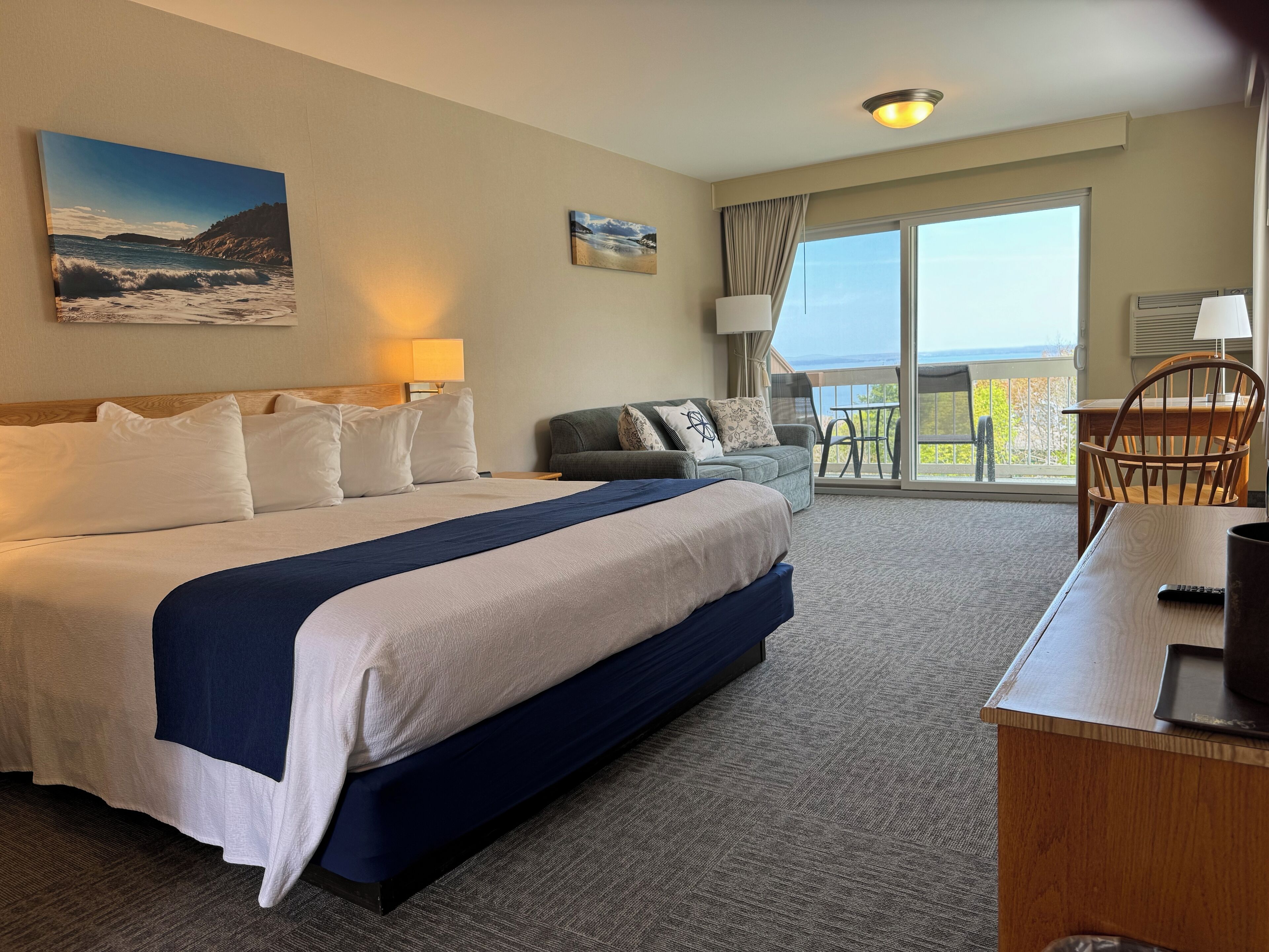 A hotel room with a large bed, a sofa, a balcony with a view, and a desk with a remote control.