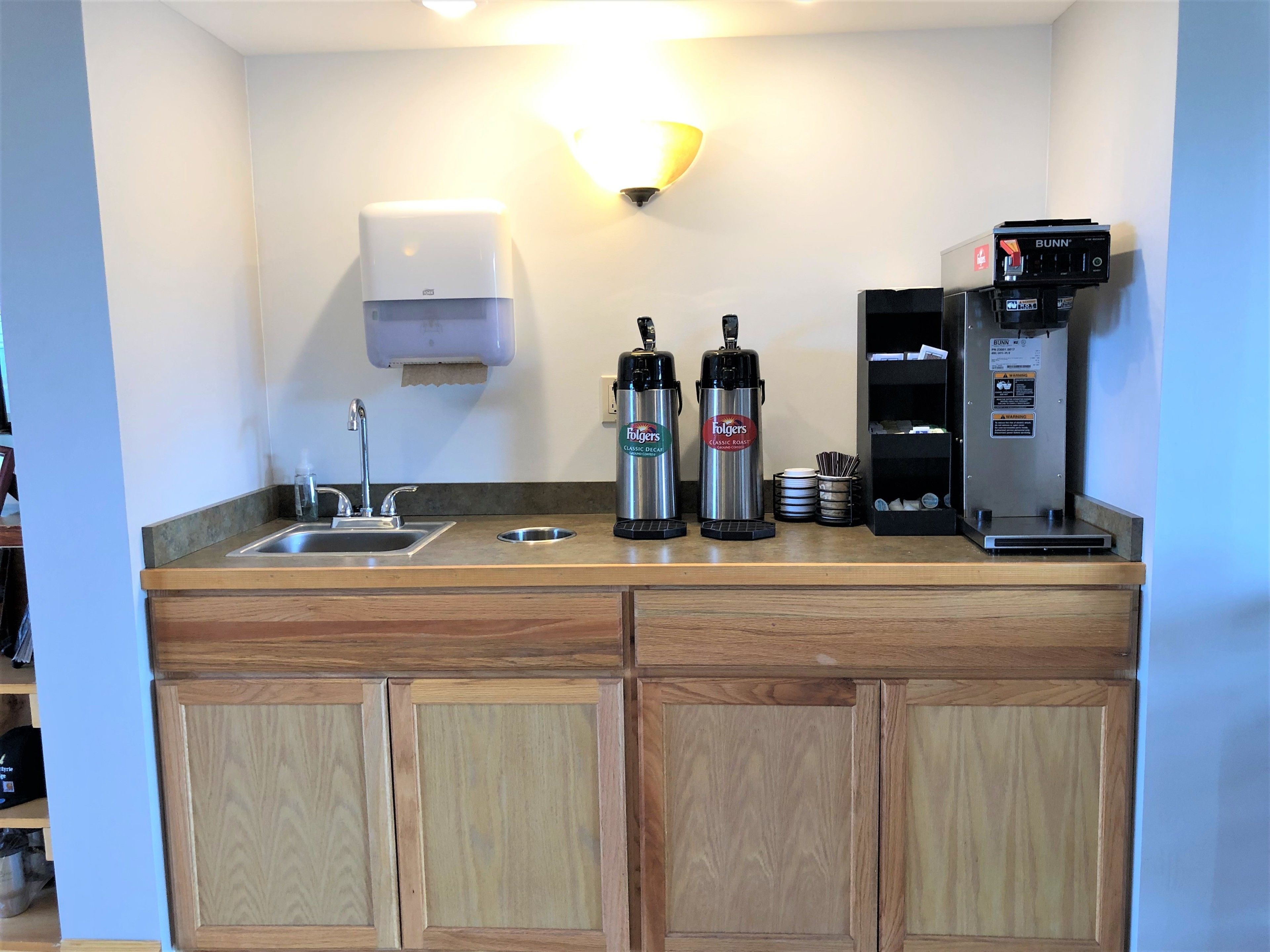 Una kitchenette con un lavabo, dos cafeteras Folgers, una máquina de café Bunn y un dispensador de toallas de papel.