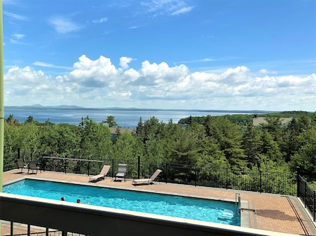 A swimming pool with lounge chairs, surrounded by a fence, overlooking a body of water and distant mountains.