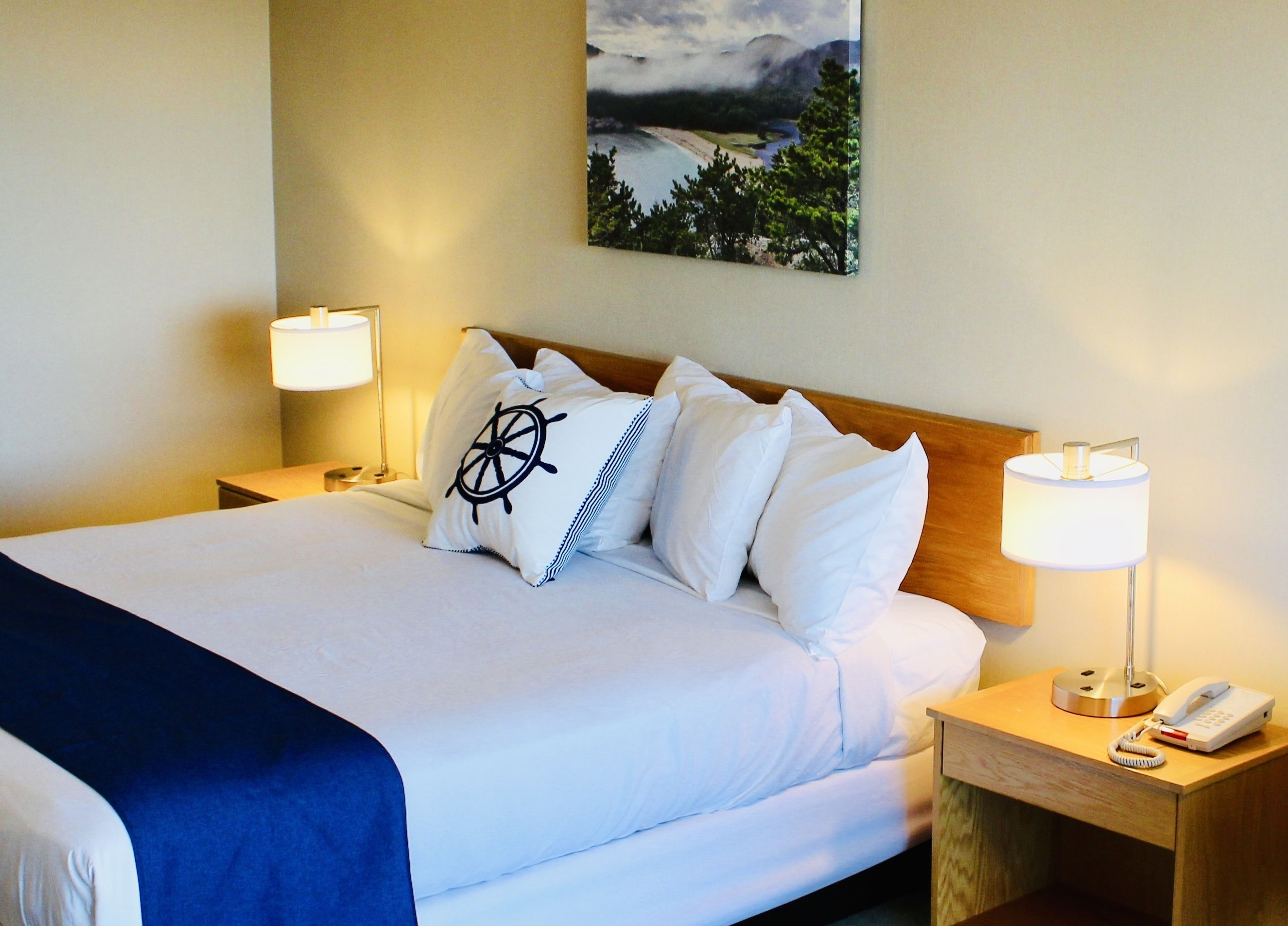 A neatly made bed with white linens and a blue throw blanket, two bedside lamps, a wooden nightstand with a phone, and a painting of a landscape on the wall.