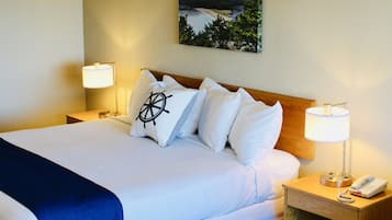 A neatly made bed with white linens and a blue throw blanket, two bedside lamps, a wooden nightstand with a phone, and a painting of a landscape on the wall.