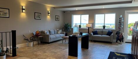 A spacious living area with two sofas, a coffee table, and a dining table. There are large windows providing a view of the outdoors.