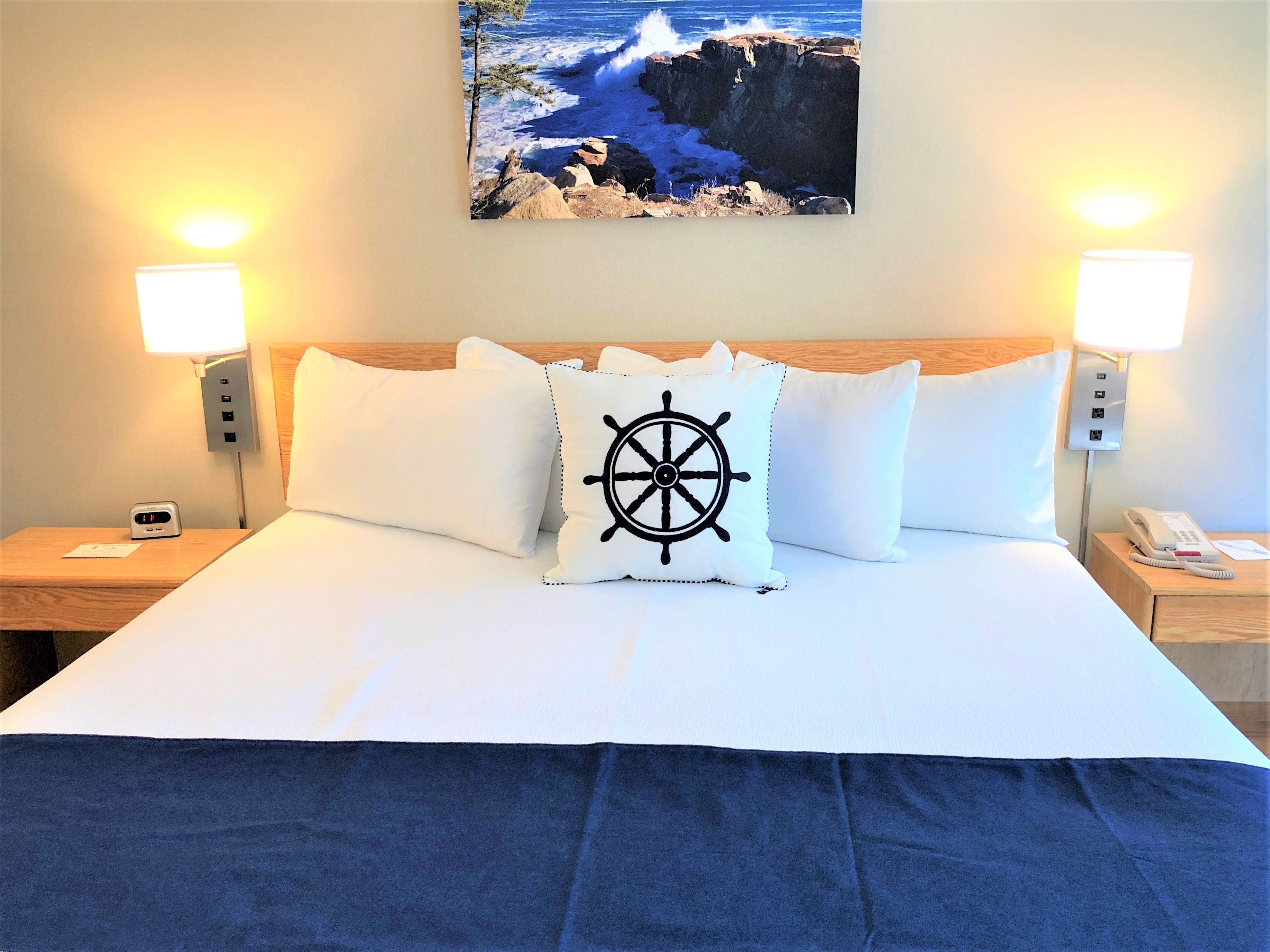 Habitación de hotel con cama, dos lámparas de noche, un reloj digital, teléfono y un cuadro de una escena costera.