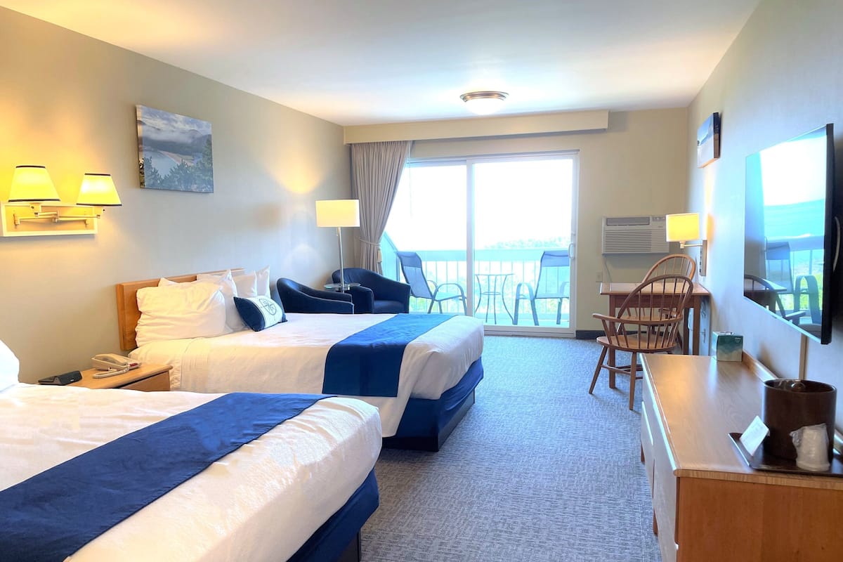 Quarto de hotel com duas camas, uma escrivaninha, cadeiras e uma varanda com vista.