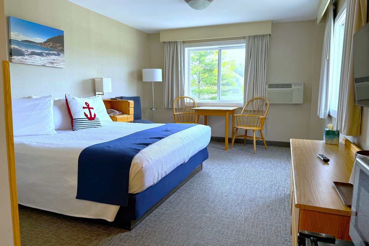 Quarto de hotel com cama, escrivaninha, cadeira, janela com cortinas, quadro na parede e ar condicionado.