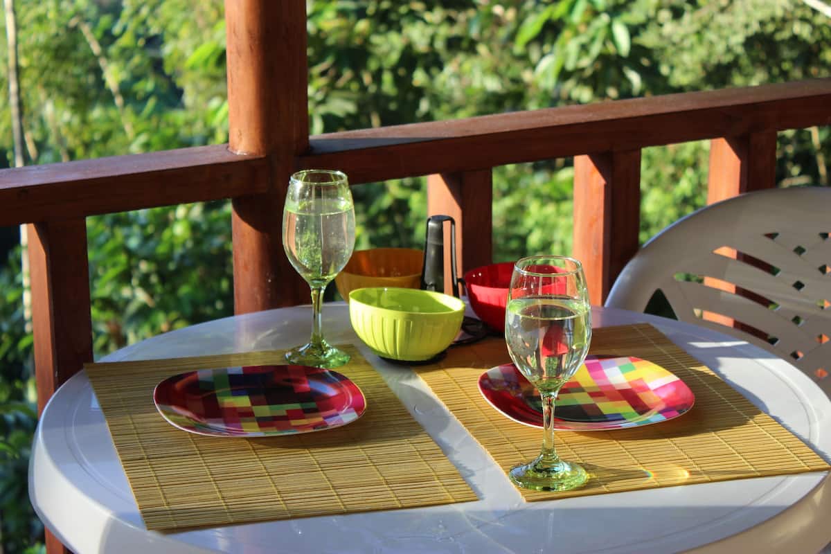 A table set with wine glasses and colorful plates on a white tablecloth, placed on a wooden deck with a view of greenery.