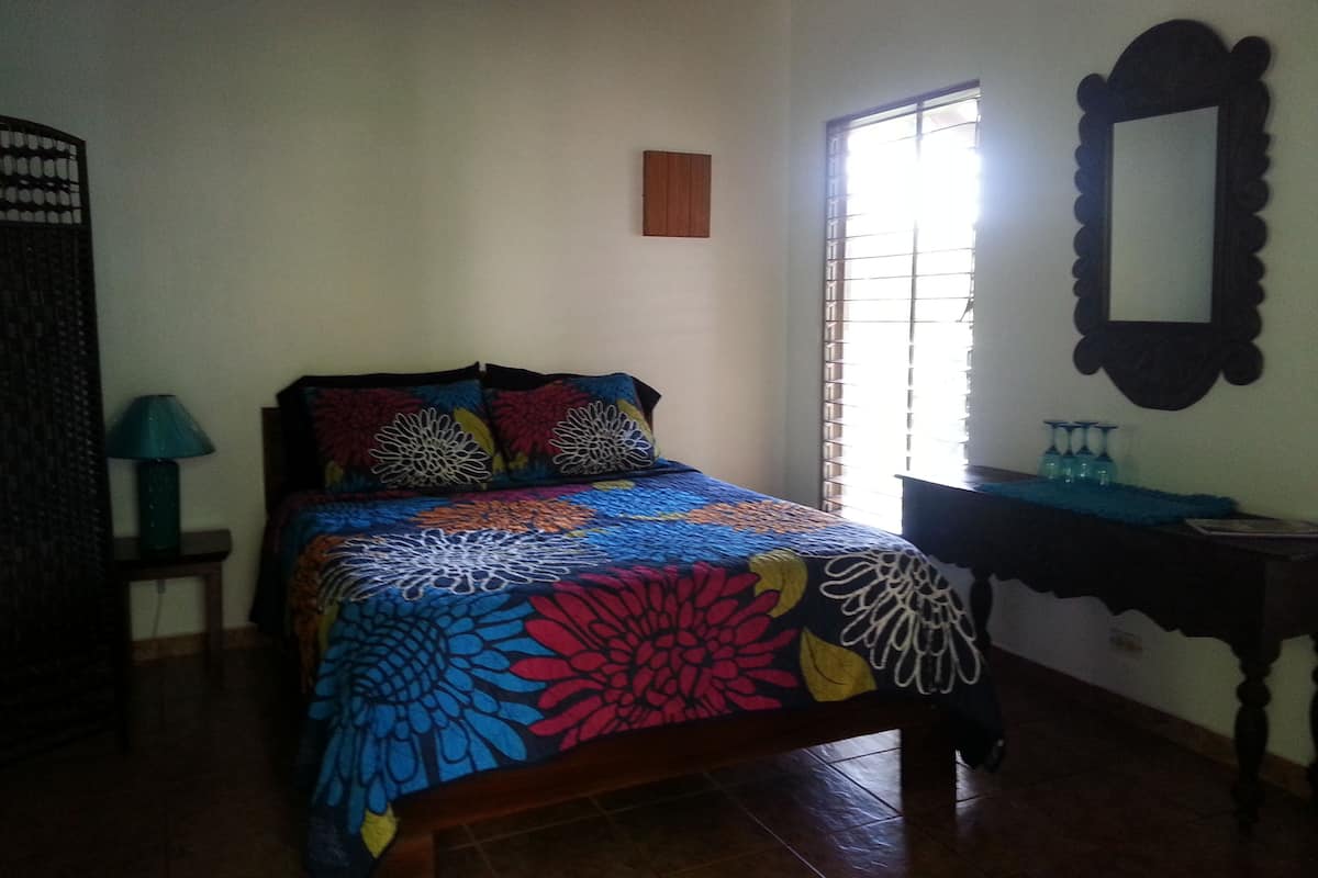 A bedroom with a bed, a nightstand, a lamp, a window with blinds, and a mirror on the wall.