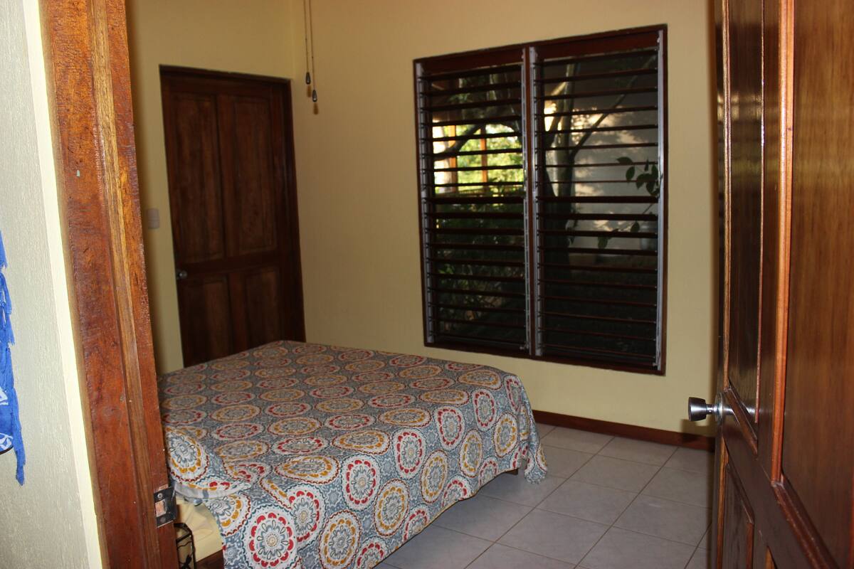 A bedroom with a patterned bedspread, a wooden door, and a window with blinds.