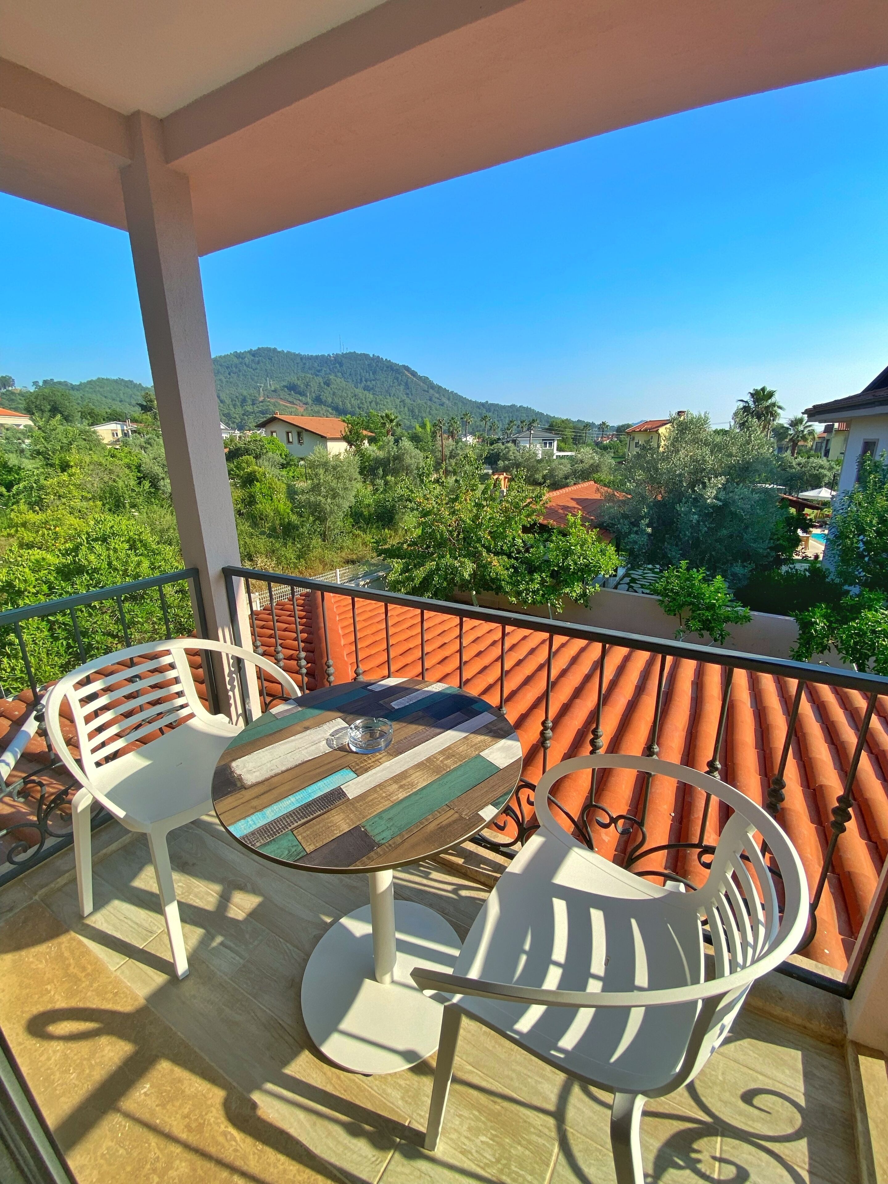 A balcony with a table and two chairs, overlooking a residential area with houses and greenery.