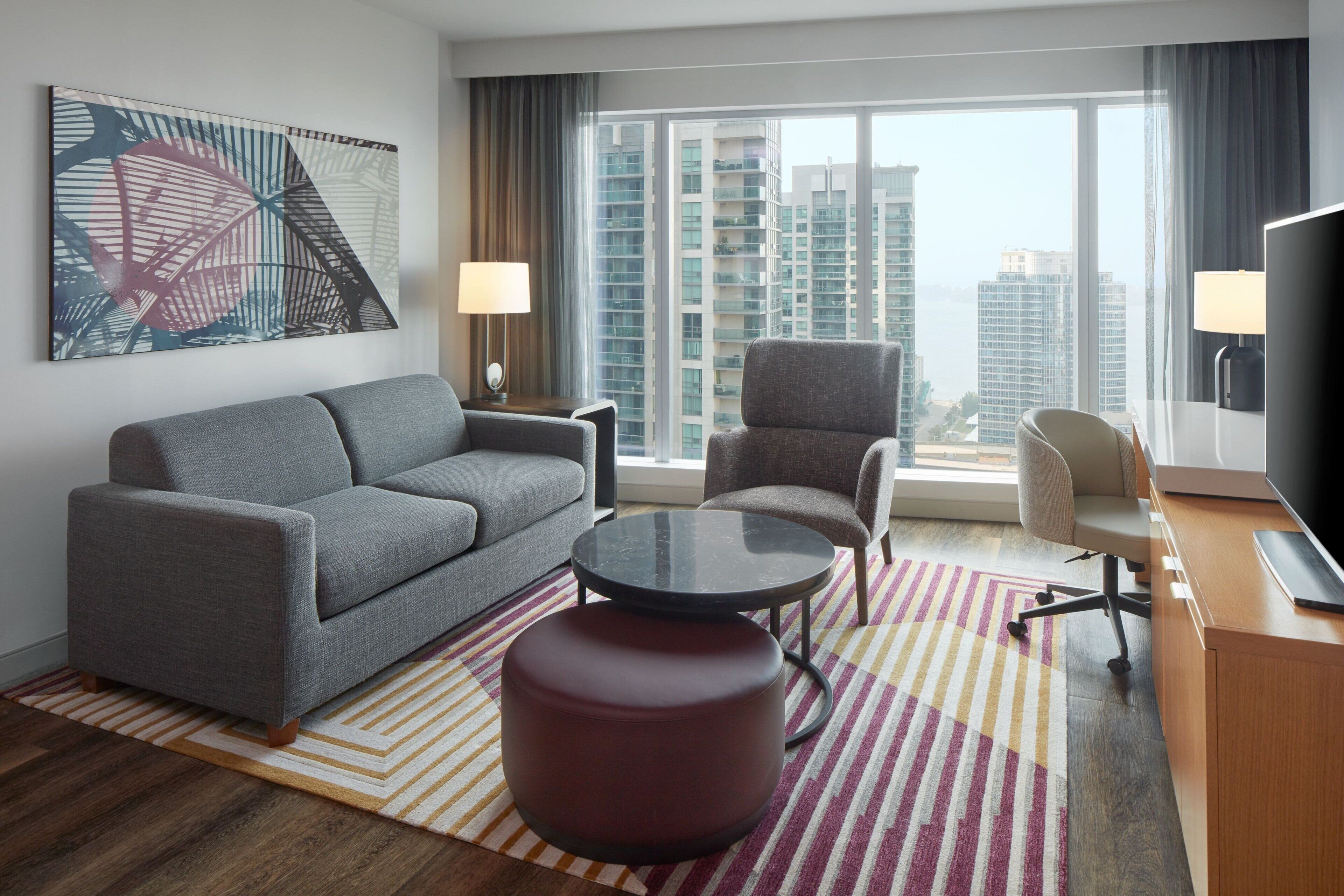 A modern hotel room with a grey sofa, a round coffee table, a red ottoman, a TV, and a large window offering a city view.
