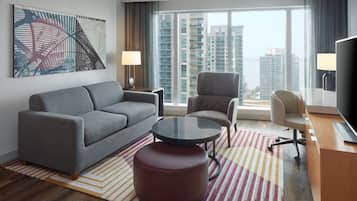 Een moderne hotelkamer met een grijze bank, een ronde salontafel, een rode poef, een televisie en een groot raam met uitzicht op de stad.